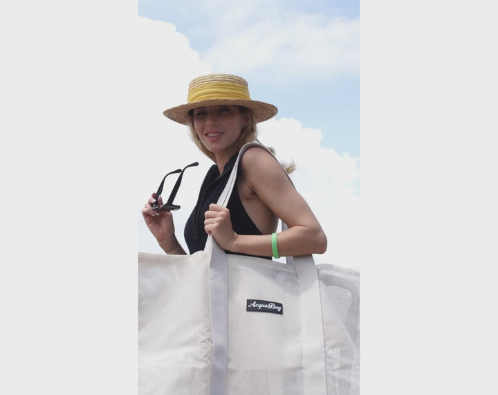 Woman wearing a straw hat and holding sunglasses, carrying an AcquaBay by ACQUABOSS mesh beach bag designed to be sand-free and spacious