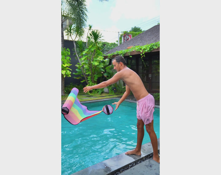 Man in pink swim shorts tossing an ACQUABOSS rainbow floating water hammock float into a pool, preparing to jump in and relax. Perfect gift for him