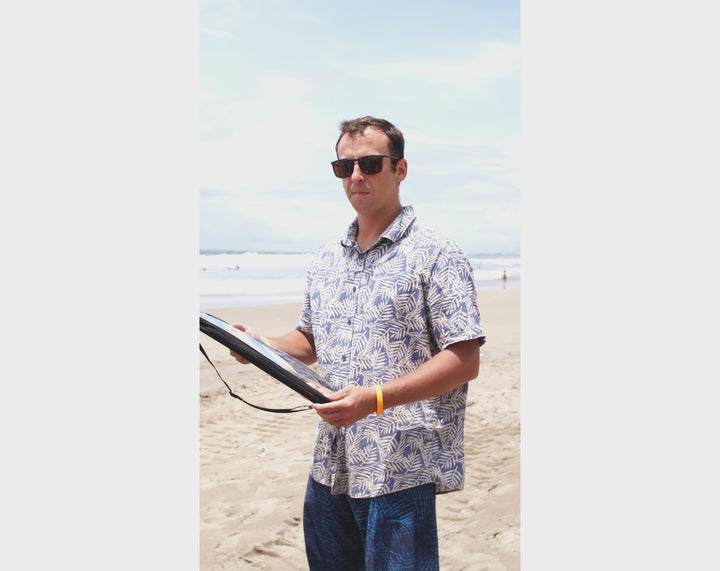 Man wearing sunglasses unboxing the ACQUABOSS Beach Sunset Paddleball Set on the sand, revealing the rainbow-coloured paddles and balls ready for a beach game