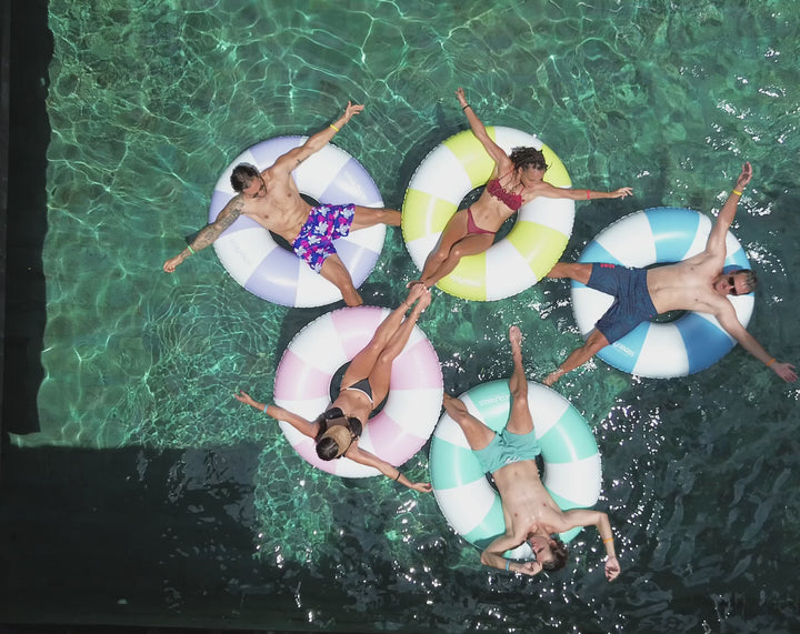 Group of friends relaxing and floating in the pool on ACQUABOSS Extra Large Drift Rings in pink, blue, yellow, purple, and green